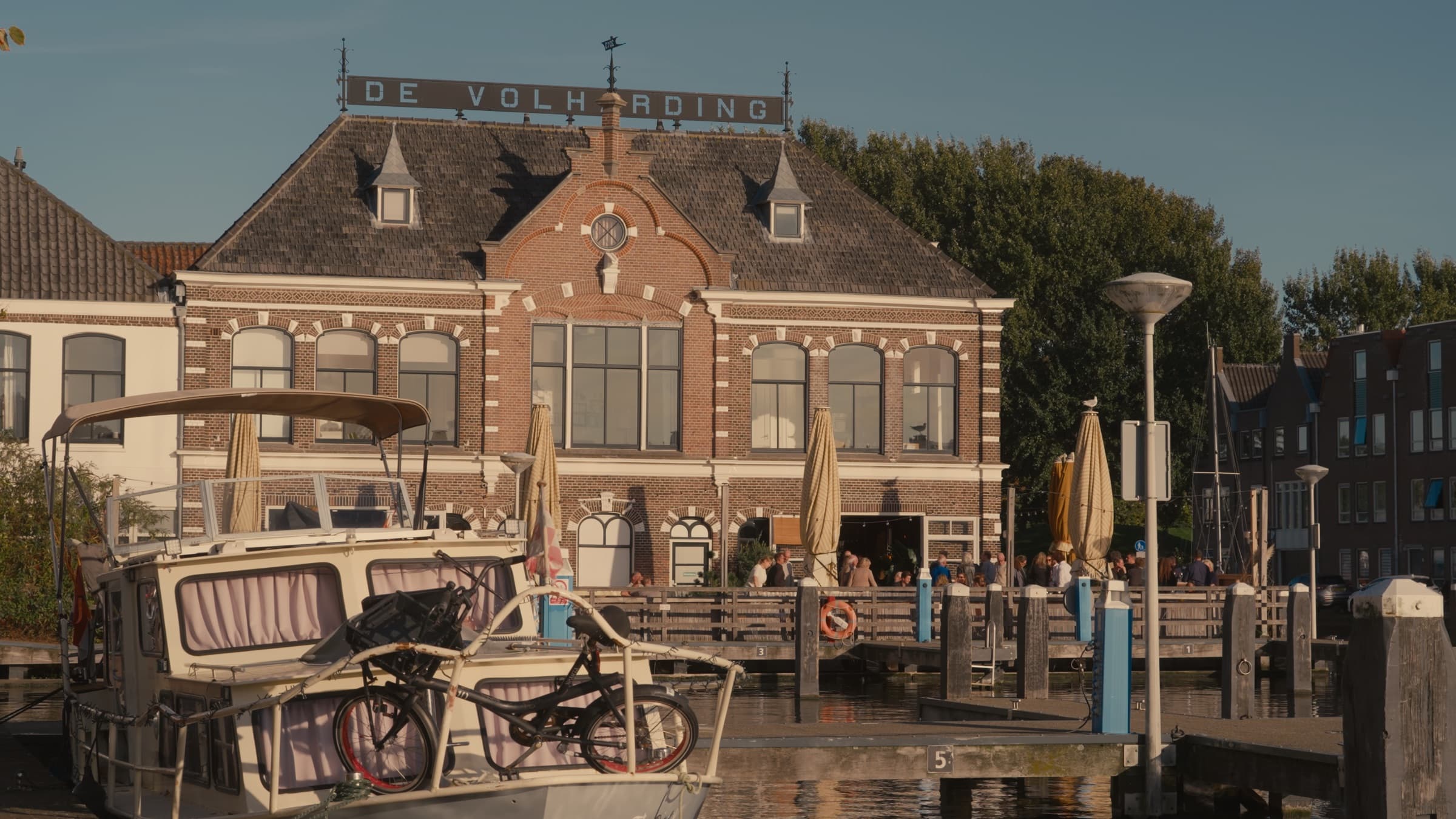 Een historisch bakstenen pand met het opschrift De Volharding aan het water, met een vol terras op de steiger en een boot met een fiets op het dek in de voorgrond, in het late middaglicht