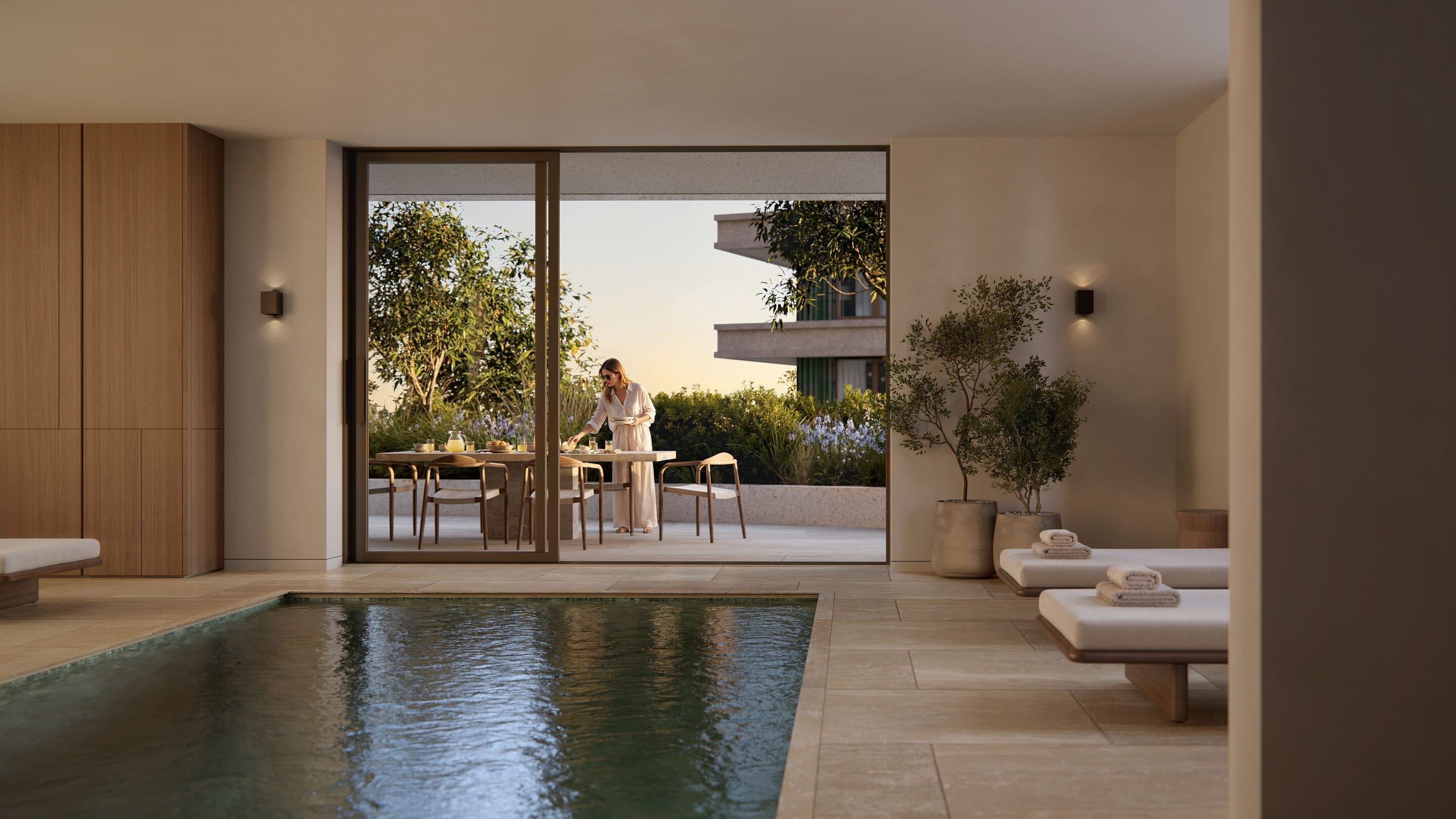 Artist impression of an indoor pool with stone floors and loungers, opening onto a planted terrace where a woman sets a table.
